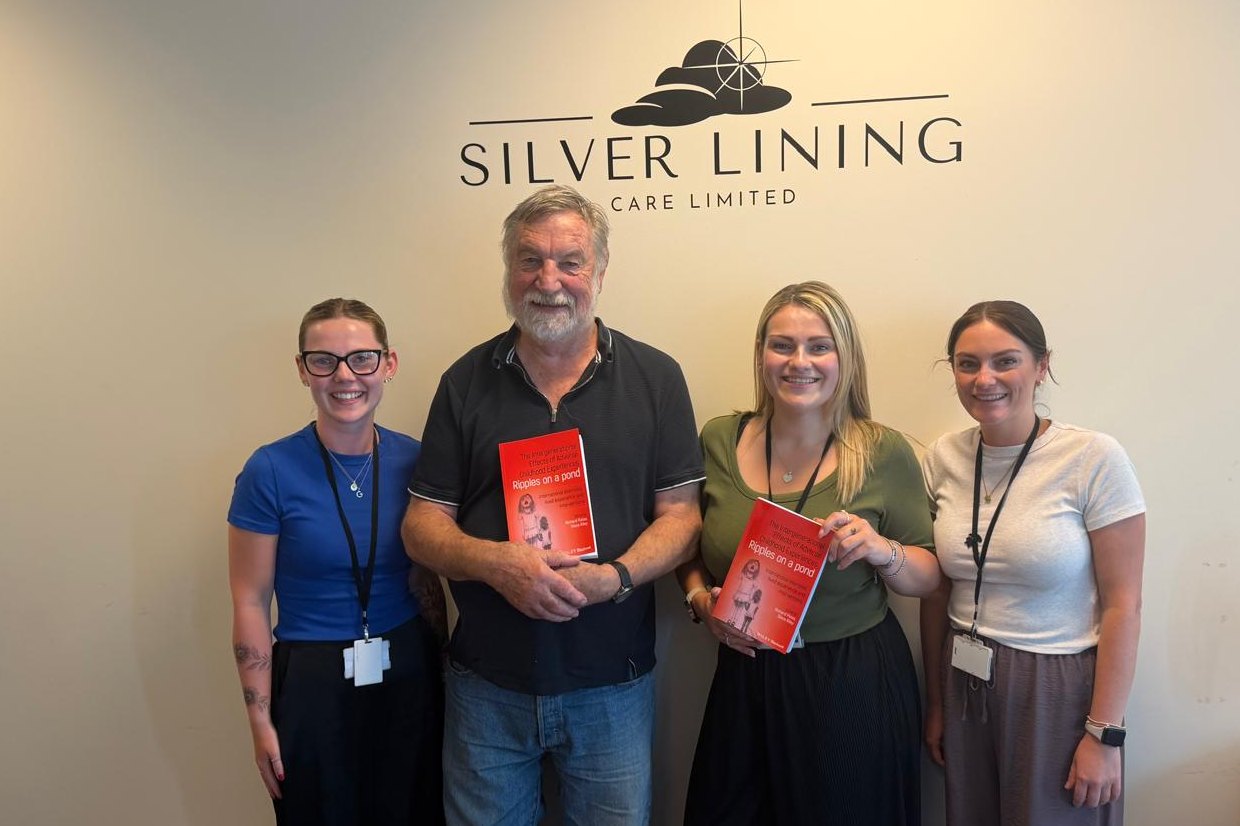 Dr Richard Pates smiling with three members of our team in front of the Silver Lining Care sign, two of them holding copies of his new book.