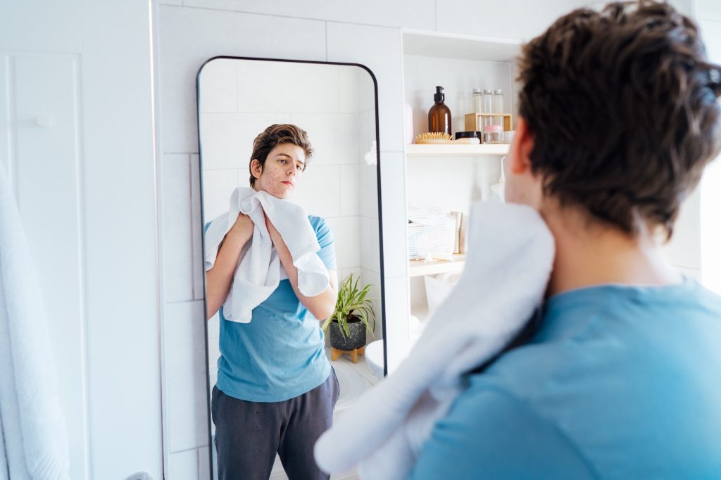 Concentrated caucasian teenage boy with acne problem takes care of his face skin at home.