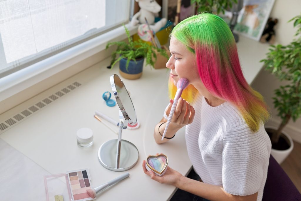 Teenager girl with dyed colored hair doing makeup looking in mirror